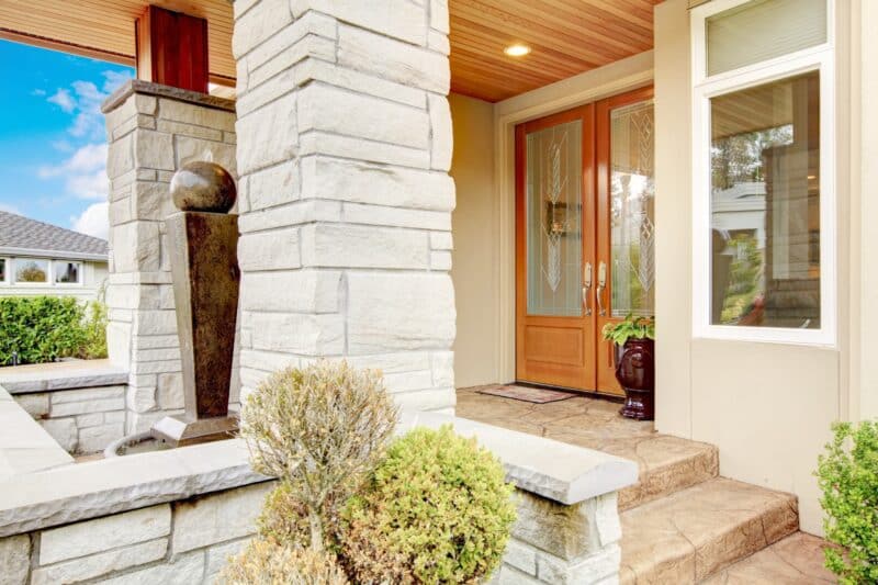 large wixom house front porch with stone columns, windows and a brown double door with large specialty windows. plants and other things surrounding the area
