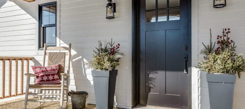 Inviting front porch with a black steel door, decorative plants in tall planters, a rocking chair with a red patterned pillow, and rustic decor elements