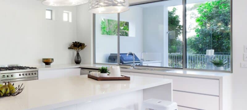 A modern kitchen with a sleek, white countertop and stainless steel appliances. Large windows provide a view of lush greenery outside, adding natural light to the space