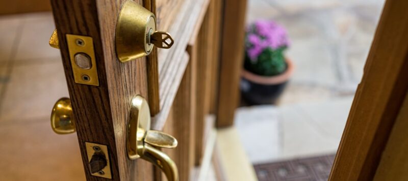 Close-up of a wooden front door slightly ajar, showing a brass deadbolt with a key inserted and a blurred flowerpot outside
